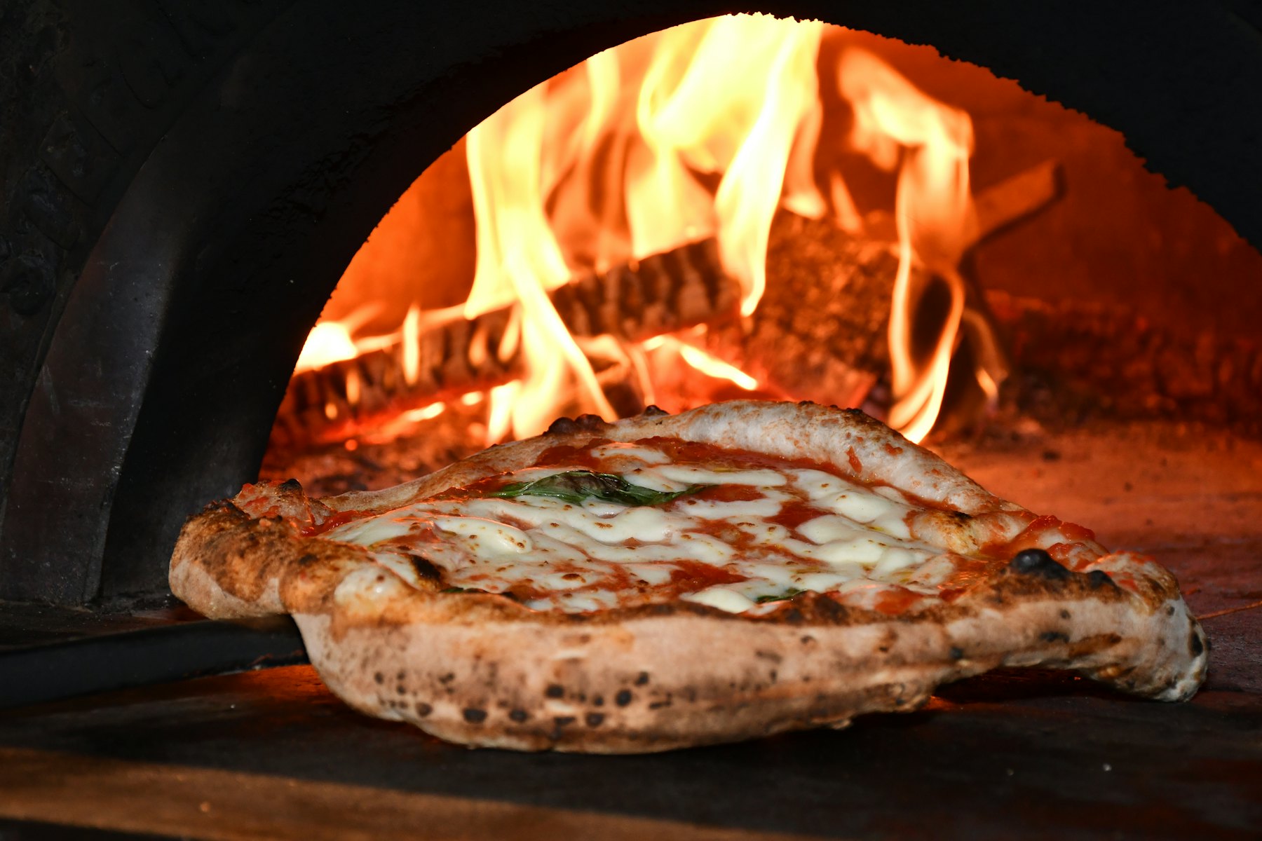 Pizza im Holzofen, dahinter lodert das Feuer